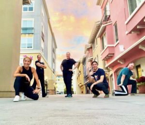 Five martial arts students in kneeling and standing poses against a blue, yellow, and pink sunset sky. The camera is angled upward and they are between pastel colored, multi-story town homes.